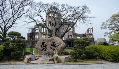 A memorial stone with Japanese characters sits in the foreground, with the skeletal remains of the Hiroshima Peace Memorial (Genbaku Dome) in the background. Bare trees and trimmed bushes surround the monument.
