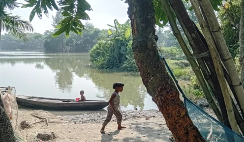 Photograph of a child walking by a lake in rural Bangladesh with a person in a boat in the background.