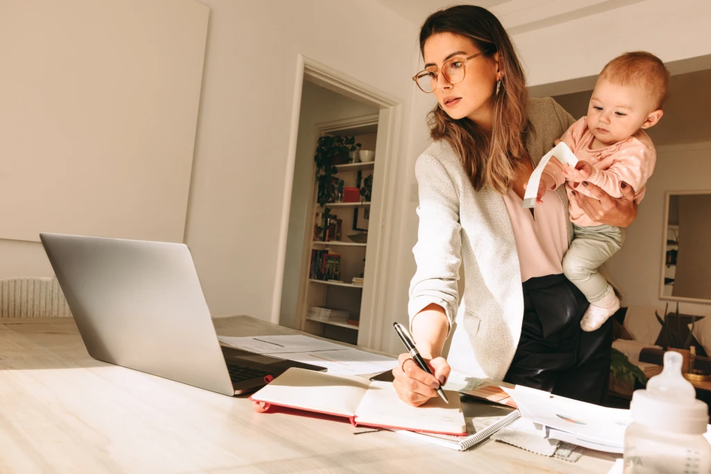 A mother multitasks, holding her child on one hip as she manages a computer and scattered workbooks.