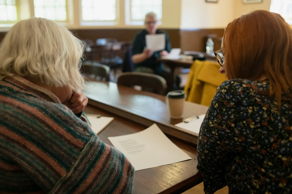 wo women are sat at a brown wooden table reading from a piece of paper. A coffee cup rests on the table. In the background a third person is reading from a page.