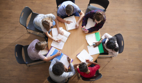 A photograph of young people sitting round a table, writing. The photograph is taken from above