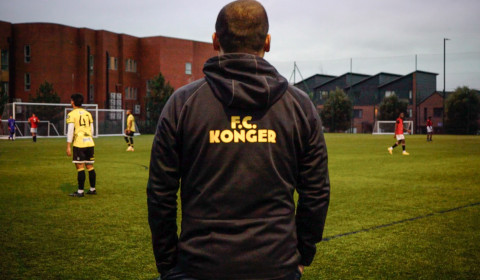 Photograph of someone watching a grassroots football match. They are wearing a Konger FC hoodie.