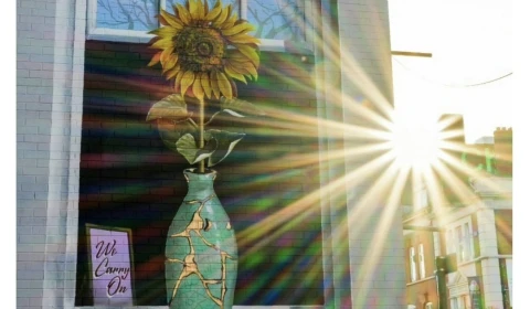 A mural of a sunflower in a kintsugi-style vase with the words “We Carry On,” lit by sunbeams, symbolizing hope and resilience.