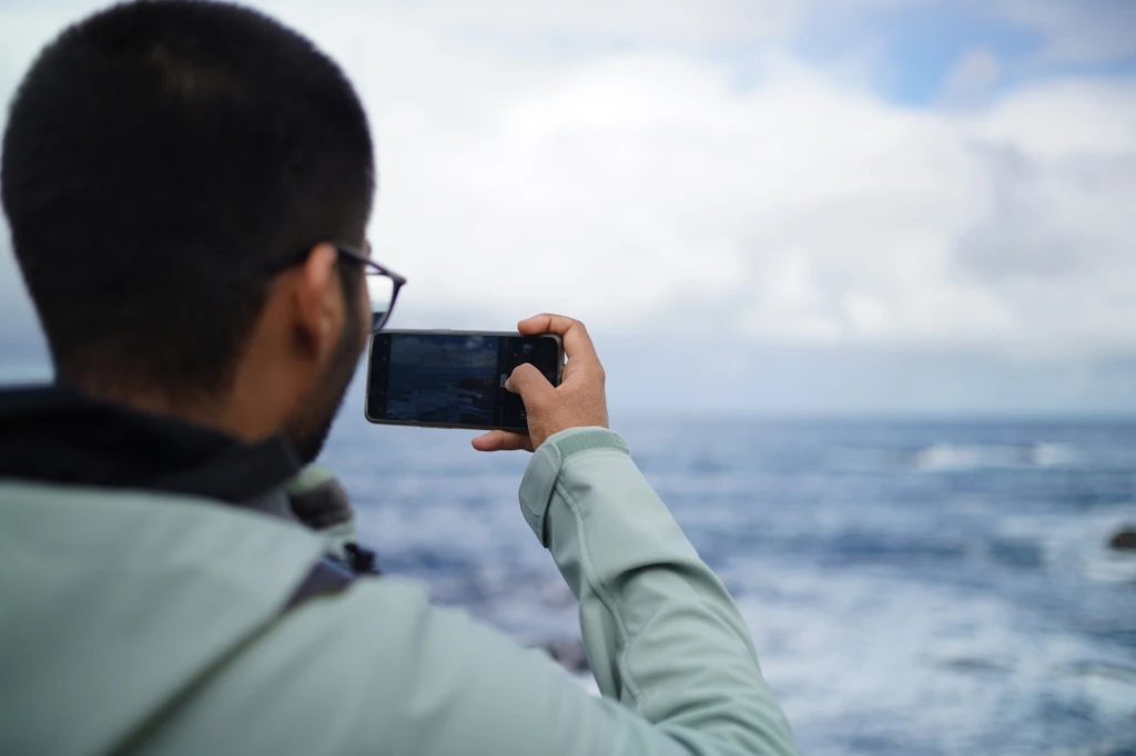 A person photographs the ocean with their phone on a cloudy day.