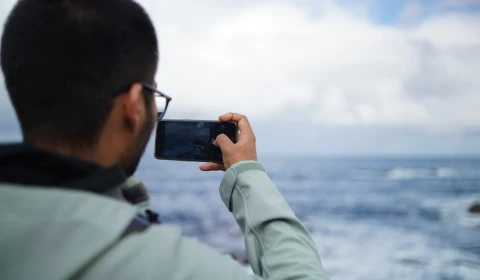 A person photographs the ocean with their phone on a cloudy day.