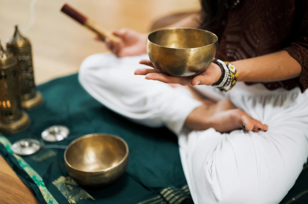 Woman wearing white trousers and a black long sleeved top is sat cross legged on a green mat.  In she is holding a small sound bowl in her left hand.