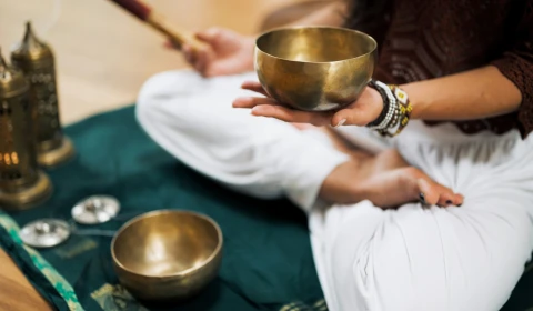 Woman wearing white trousers and a black long sleeved top is sat cross legged on a green mat.  In she is holding a small sound bowl in her left hand.