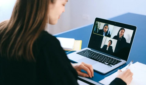 An image of a girl on a video call with three other team members. She is holding a pen to paper.