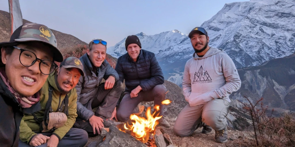 Photograph of a group of individuals around a camp fire in the mountains, with a snow covered mountain in the background