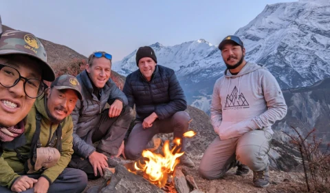 Photograph of a group of individuals around a camp fire in the mountains, with a snow covered mountain in the background
